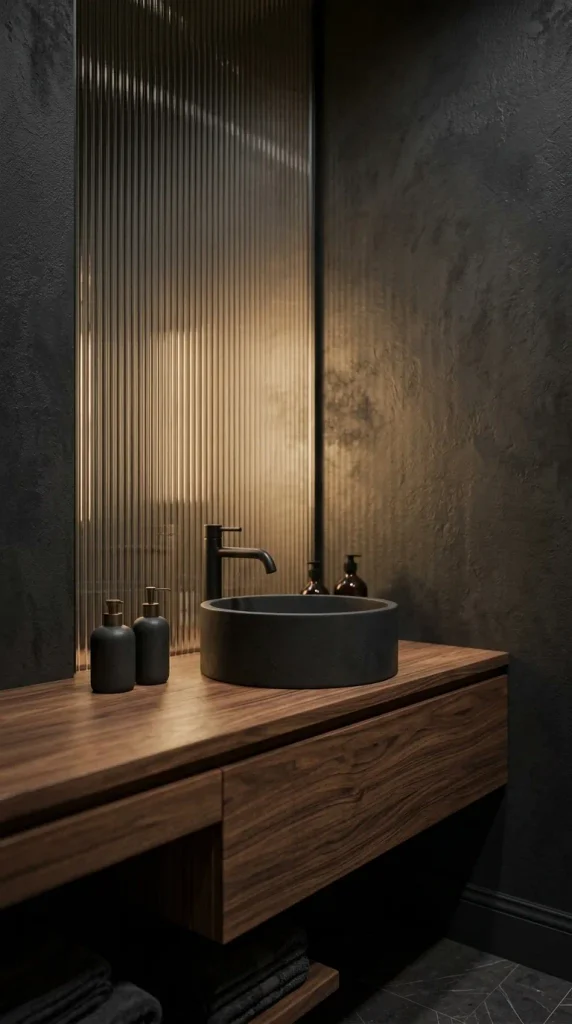 A close-up view of a floating walnut wood bathroom vanity with a black cylindrical concrete vessel sink, matte black faucet, and three dark soap bottles, set against textured charcoal walls with reeded glass.